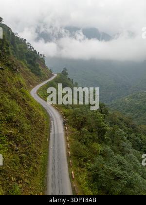 Eine schmale Bergstraße kurvt entlang eines steilen Hügels durch dichten Wald, mit Nebel und niedrigen Wolken, die über den umliegenden Tälern hängen. Stockfoto
