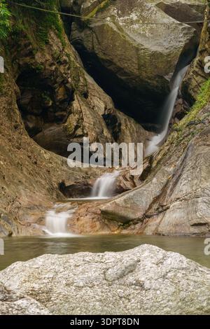 Ein kleiner Wasserfall fließt über glatte Felsformationen in einen flachen Pool, umgeben von Naturstein und Vegetation. Stockfoto