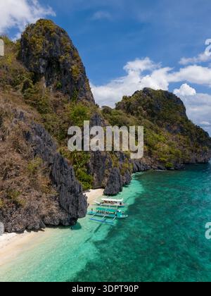 Ein Blick aus der Vogelperspektive zeigt steile Kalksteinklippen, die an einen schmalen Sandstrand und flaches türkisfarbenes Wasser grenzt, mit Auslegerbooten, die in der Nähe der Küste ankern. Stockfoto