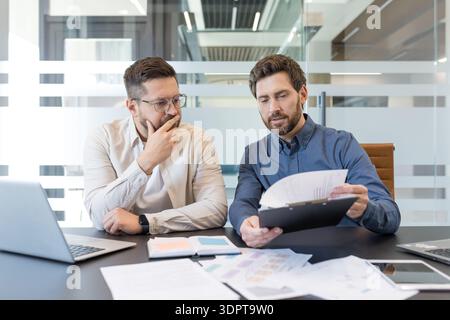 Erfahrene Geschäftsleute analysieren die Finanzberichte des Unternehmens, prüfen wichtige Dokumente und diskutieren einen neuen Projektvertrag während eines offiziellen Geschäftstreffens in einem modernen Büro Stockfoto