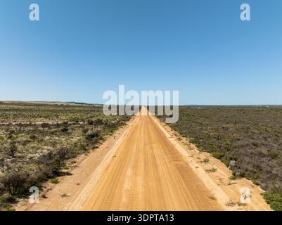 Ein erhöhter Blick aus der Luft auf eine gerade, unbefestigte Straße, die sich durch offenes Buschland und niedrige Vegetation unter klarem blauem Himmel erstreckt. Stockfoto