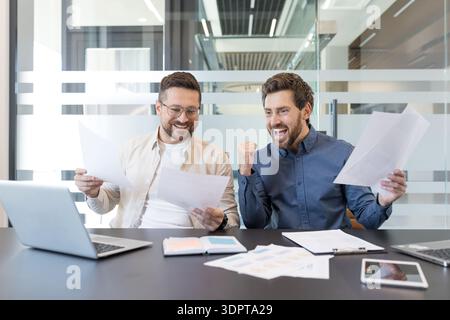 Zwei glückliche männliche Geschäftspartner feiern gemeinsam im modernen Büro Erfolge und finanziellen Erfolg und sehen sich die Dokumente mit Spannung und Erfolg an Stockfoto