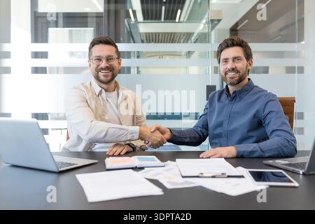 Zwei lächelnde männliche Geschäftspartner sitzen an einem modernen Bürotisch, schütteln die Hände, bestätigen eine Partnerschaft oder einen erfolgreichen Abschluss nach einem produktiven Meeting Stockfoto