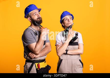 Bauarbeiter stehen tief in Gedanken, mit Werkzeugen, die unter dem Kinn liegen, und entwickeln Ideen für Bauprojekte. Männliche und weibliche Renoviererinnen arbeiten bei der Problemlösung von Renovierungsproblemen zusammen. Stockfoto