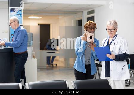 Eine ältere Ärztin spricht mit einer jungen Frau, die im Wartebereich der Klinik Halsband trägt. Der afroamerikanische Patient hört aufmerksam zu, während der Arzt mit dem Klemmbrett die sanften Erholungsmöglichkeiten erklärt. Stockfoto