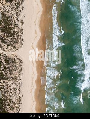 Ein Blick von oben nach unten zeigt einen Sandstrand, der auf Meereswellen trifft, mit Küstenvegetation und einem schmalen Pfad, der zur Küste führt. Stockfoto