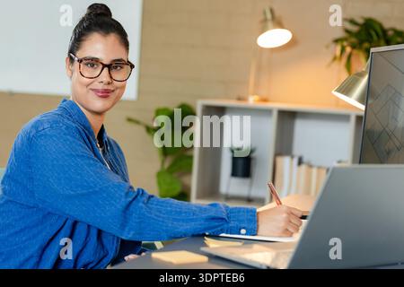 Frau mit blauem Hemd, die Notizen im Heimbüro mit Laptop, Lampe und Pflanzen schreibt Stockfoto
