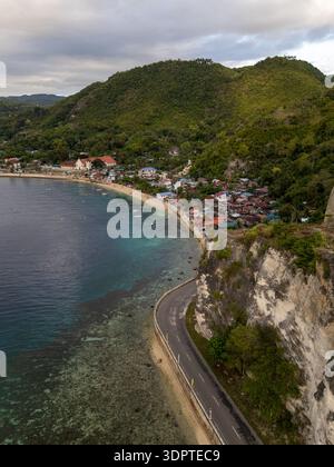 Eine Küstenstraße führt entlang steiler Klippen neben klarem Meereswasser, mit einer kleinen Küstenstadt und bewaldeten Hügeln im Hintergrund. Stockfoto