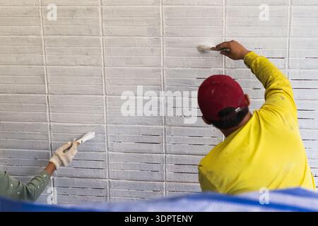 Zwei Bauarbeiter, die während eines Renovierungsprojekts weiße Farbe mit Pinseln auf eine strukturierte Ziegelwand auftragen. Stockfoto