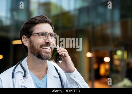 Lächelnder männlicher Arzt im Laborkittel und Stethoskop spricht auf Handy vor einer modernen Klinik, vermittelt Telemedizin, Gesundheitskommunikation, Professionalität und Patientenbetreuung Stockfoto