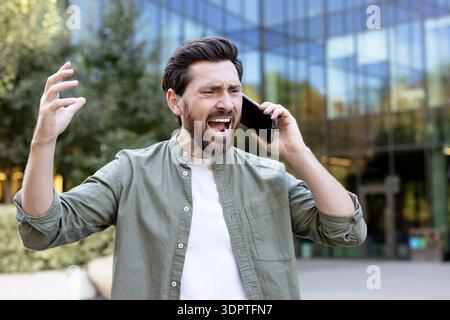Der Mann fühlt sich frustriert und gestresst, drückt Wut und Unzufriedenheit aus, während er draußen mit einem Mobiltelefon kommuniziert, mit einer ausdrucksstarken, emotionalen Reaktion Stockfoto