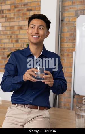 Asiatischer Mann sitzt auf hölzernem Tischrand im Büro, hält ein Glas Wasser und lächelt Stockfoto