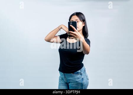 Eine junge asiatische Frau in einem schwarzen T-Shirt, die mit ihrem Smartphone ein Selfie macht oder Vloggen macht und ihr Gesicht teilweise vor weißem Hintergrund verdeckt. Stockfoto