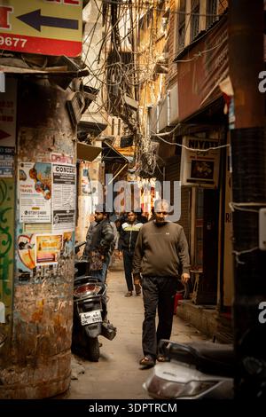 Männer gehen durch eine enge, sonnendurchflutete Gasse voller verworrener Drähte und Plakate und fangen die chaotische Energie des täglichen Lebens in Varanasi, Indien, ein. Stockfoto