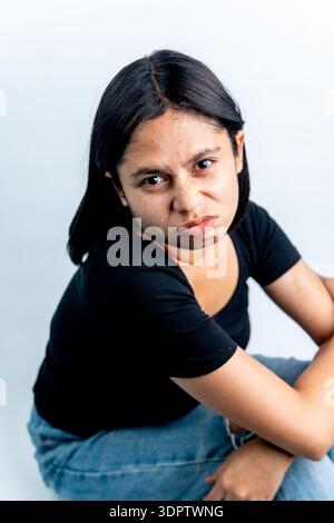 Intensives Porträt einer südostasiatischen Frau in schwarzem T-Shirt und Jeans, die auf dem Boden sitzt. Isoliertes Studio, aufgenommen auf weiß, Konzept von Fokus oder Trauer. Stockfoto