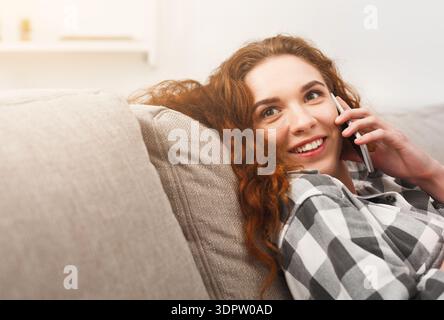 Junge Frau, die am Telefon spricht, während sie auf einer Couch in einem hellen Wohnzimmer sitzt Stockfoto