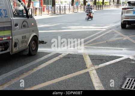 Straßenszene mit Nutzfahrzeug, das Wasser auf Asphaltstraße sprüht, städtische Kreuzung in China, Fußgänger und Roller im Hintergrund Stockfoto
