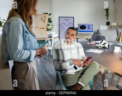 Verschiedene Kollegen diskutieren Smartphone-Daten per Laptop mit VR-Headset und rosa Kopfhörern im Büro Stockfoto