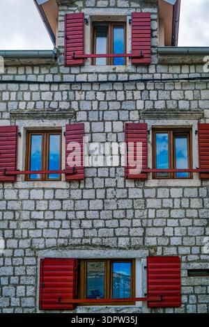 Aus nächster Nähe ein Steingebäude mit roten Holzläden und Fenstern. Die Struktur hat ein rustikales Design und ist vor einem bewölkten Himmel gesetzt. Stockfoto