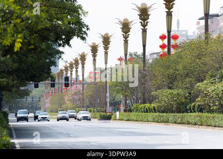 Breite Stadtstraße in China mit palmenförmigen Ampeln, roten Laternen, grünen Bäumen und Autos, die unter bewölktem Himmel fahren Stockfoto