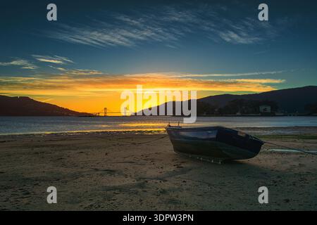 Holzboot am Strand bei goldenem Sonnenuntergang mit Rande Bridge Silhouette, Cesantes Beach, Ría de Vigo, Spanien Stockfoto