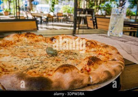 Eine frisch gebackene Pizza mit goldener Kruste und geschmolzenem Käse steht auf einem Holztisch. Ein Glas Limonade befindet sich in der Nähe in einer zwanglosen Atmosphäre. Stockfoto