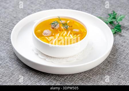 Ein heller, von oben nach unten geschnittener Schuss einer klaren, goldenen Hühnerbrühe mit kleinen Fleisch- oder Hühnerfleischbällchen, dünnen Einudeln und Karottenstücken in einer weißen Schüssel Stockfoto