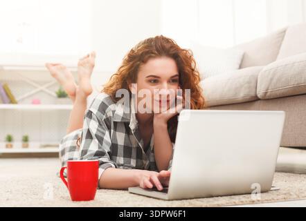 Frau, die Laptop benutzt, während sie auf Teppich im Wohnzimmer liegend, mit Kaffeetasse in der Nähe Stockfoto