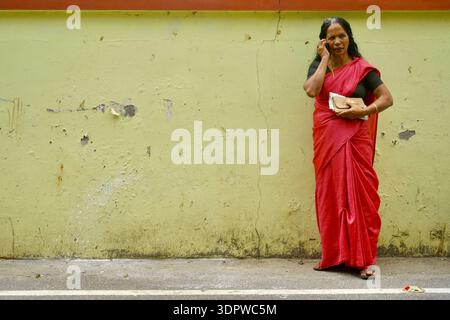 Ältere indische Frau in rotem Saree, die auf dem Handy spricht, steht an der bunten Wand in Alappuzha, Kerala, Indien Stockfoto