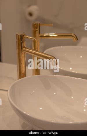Nahaufnahme eines modernen Luxusbadezimmers mit weißem Keramikwaschbecken und elegantem goldenem Wasserhahn mit sanfter, warmer Beleuchtung. Reinigen Sie minimale Innenräume Stockfoto