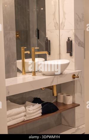 Luxuriöses, modernes Badezimmer mit weißem Waschbecken, goldenem Wasserhahn und Marmorfliesen. Saubere Handtücher und Toilettenpapier auf offenen Holzflächen gelagert Stockfoto