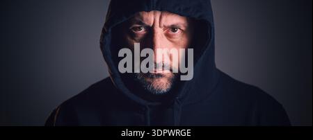 Porträt eines Mannes mittleren Alters mit kurzem Bart und verwittertem Gesicht, der einen dunklen Hoodie trägt und direkt mit gefurchten Augenbrauen und intensivem Ga starrt Stockfoto