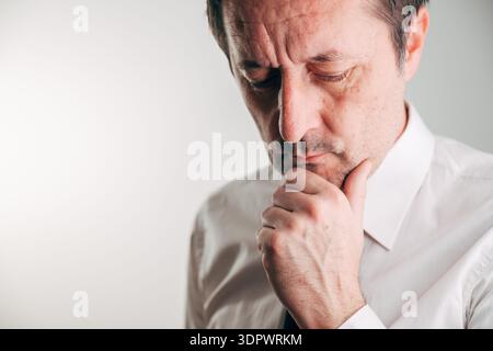Besorgter Geschäftsmann mittleren Alters in weißem Hemd und Krawatte, der mit der Hand am Kinn nach unten blickt, Stress, Sorge und tiefe Gedanken auf einem neutralen Rücken ausdrückt Stockfoto