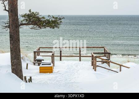Schneebedeckter Aussichtspunkt am Meer mit Holzbänken mit Blick auf die ruhige Wintersee, die eine friedliche und minimale Küstenlandschaft schafft Stockfoto