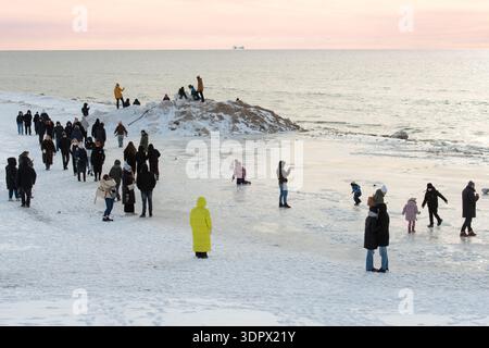 Eisige Winterlandschaft mit gefrorenen Wellen entlang einer verschneiten Küste und Menschen, die auf der Ostsee in Palanga, Litauen, spazieren gehen Stockfoto