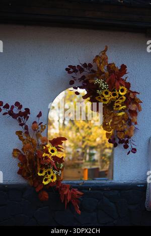 Ein Herbstkranz aus bunten Herbstblättern und hellgelben Blumen ziert einen Fensterrahmen und schafft eine gemütliche saisonale Atmosphäre mit warmen Tönen Stockfoto
