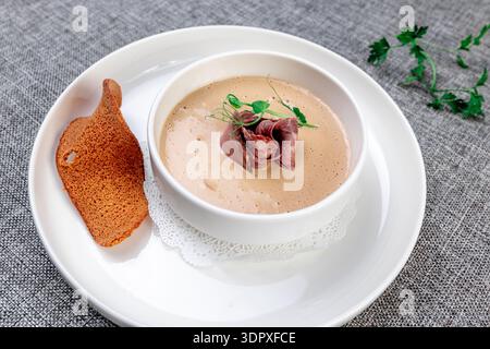 Eine reichhaltige und cremige Wildpilzsuppe (wahrscheinlich Porcini oder Pfifferlinge), elegant in einer weißen Schüssel auf einem großen Präsentationsteller präsentiert. Die Suppe ist g Stockfoto