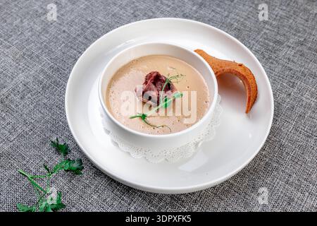 Eine reichhaltige und cremige Wildpilzsuppe (wahrscheinlich Porcini oder Pfifferlinge), elegant in einer weißen Schüssel auf einem großen Präsentationsteller präsentiert. Die Suppe ist g Stockfoto