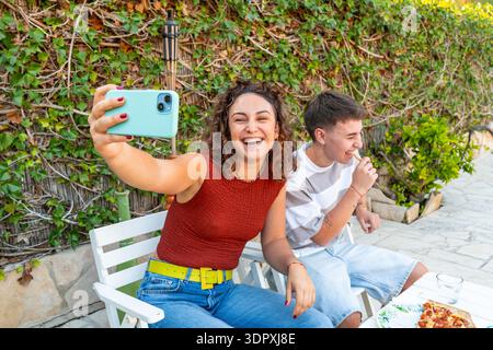 Junge Frau, die ein Selfie macht, während sie mit einem Transgender auf einer sonnigen Terrasse sitzt. Entspannte Szene mit Freundschaft, Pizza und e Stockfoto