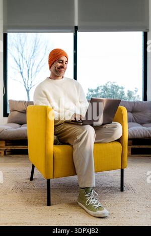 Eine entspannte Person sitzt bequem auf einem gelben Stuhl und tippt auf einem Laptop, mit einer leuchtend orangefarbenen Mütze und neutraler Kleidung, was eine warme und gemütliche Atmosphäre schafft Stockfoto