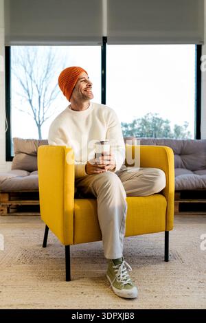 Lächelnder Mann in einem weißen Pullover und orangefarbenen Beanie, der Kaffee genießt, während er in einem modernen gelben Stuhl in einem gemütlichen Wohnzimmer mit großen Fenstern und n sitzt Stockfoto