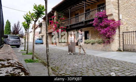 Ein paar spazieren Hand in Hand durch eine kopfsteingepflasterte Straße im bezaubernden Colombres, Spanien, umgeben von historischer Architektur und lebendigen Blumen Stockfoto