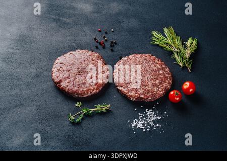 Zwei rohe Kalbsburger-Patties werden auf dunkler Oberfläche gelegt, begleitet von frischem Rosmarin, Thymian, Kirschtomaten und grobem Salz. Perfekt zum Grillen oder Stockfoto