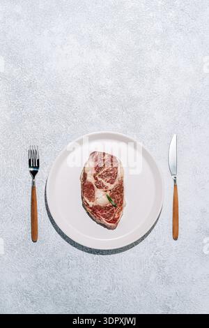 Ein erstklassiges, nasses Ribeye-Steak aus Wagyu-Rindfleisch, auf einem weißen Teller mit eleganten Essutensilien. Die marmorierte Textur unterstreicht den reichen Fl Stockfoto