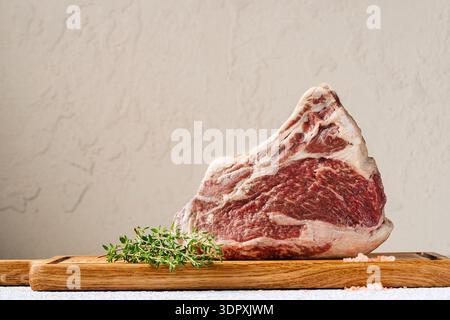 Ein rohes Ribeye-Steak auf einem Holzbrett, ergänzt durch frischen Thymian und grobe rosa Salzkristalle. Die Marmorierung des Steaks unterstreicht seine reiche Vielfalt Stockfoto