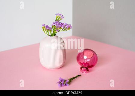 Ein minimalistisches Arrangement mit einer weißen Keramikvase mit lila Blumen und einer rosafarbenen Glasvase auf der Seite auf einem weichen Hintergrund. Perfekt für Stockfoto