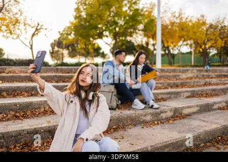 Studenten sitzen im Herbst auf Stufen in einem Park und genießen eine Lernpause. Ein Schüler macht ein Selfie, während andere sich unterhalten und lesen Stockfoto