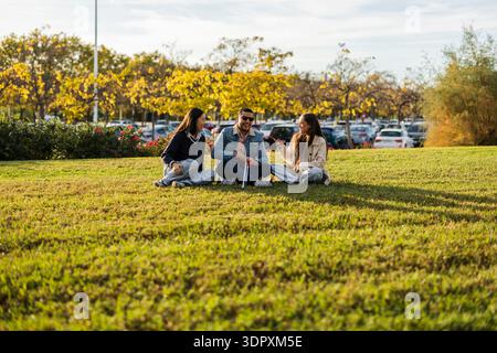 Drei Freunde sitzen auf dem Gras in einem Park, lachen und plaudern. Man verwendet einen weißen Stock, der Inklusion und Vielfalt symbolisiert. Die Sonne scheint hell o Stockfoto