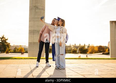 Drei Freunde feiern den Abschluss mit einem fröhlichen Selfie im Freien. Der Student im Zentrum trägt eine Mortarboard, die ihr akademisches Achie symbolisiert Stockfoto