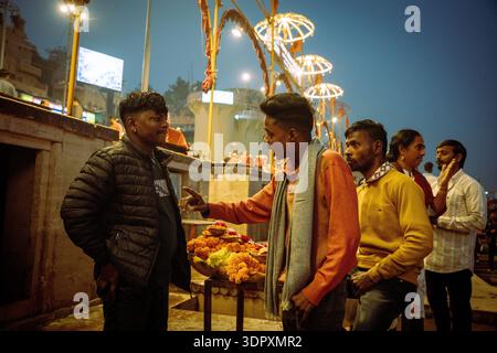 Männer sprechen in der Nähe von Blumenopfern auf einer Straße in Varanasi, Indien, während der Abendstunden mit festlichen Lichtern. Stockfoto